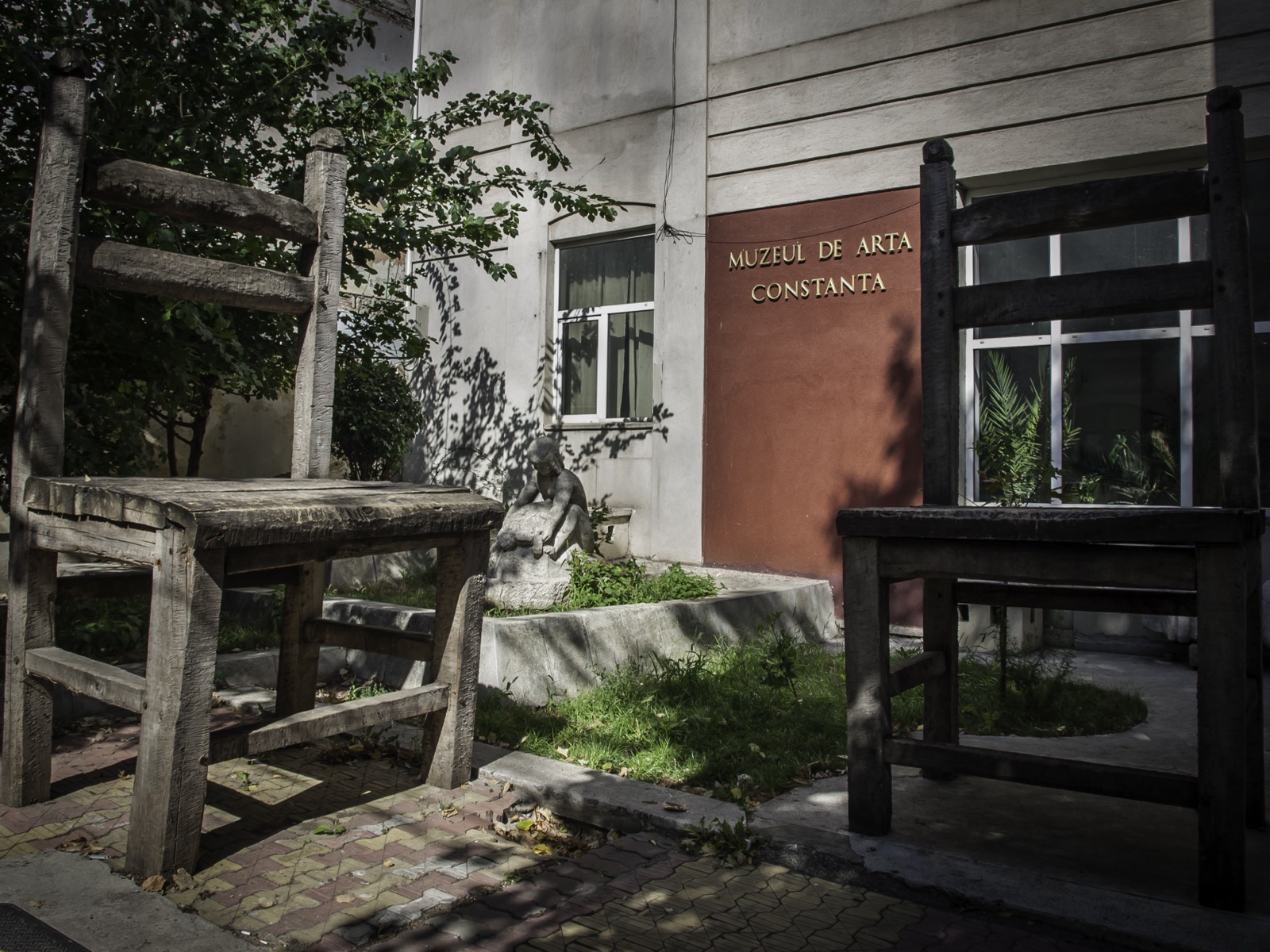 The entrance with the “Muzeul de Artă Constanța” lettering, flanked by two giant wooden chairs, with a small sculpture in the courtyard