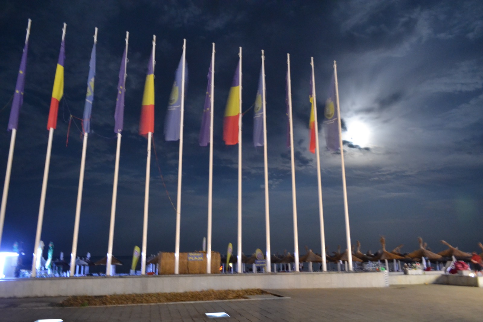 Șirul de steaguri de pe promenada plajei La Steaguri, noaptea, cu luna printre nori